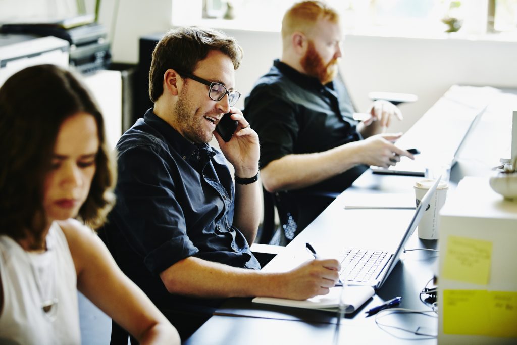 Smiling businessman sitting at workstation in startup office talking on smartphone while taking notes on notepad